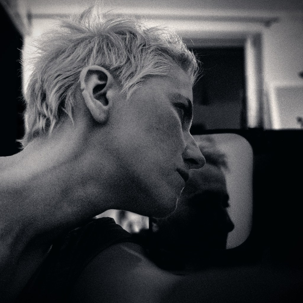 Black and white close-up portrait photo of a short-haired person (Udnua) in side profile, leaning forward in soft indoor light with a moody, introspective atmosphere.