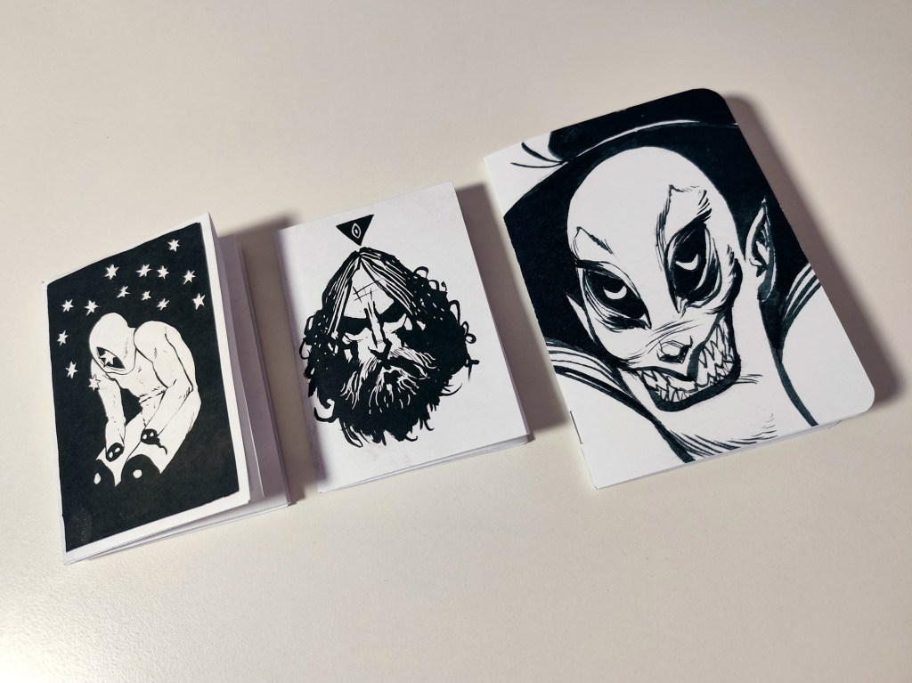 Photo of three small surreal black-and-white zines on a table, each featuring an ink illustration on the cover (a crouched figure under stars, a bearded face, and a grinning masked character).
